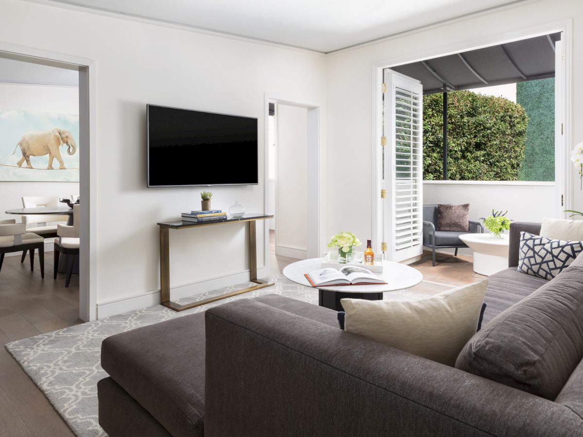 A modern living room with a gray couch, TV, artwork, and dining area. There's an open door leading to an outdoor patio space.