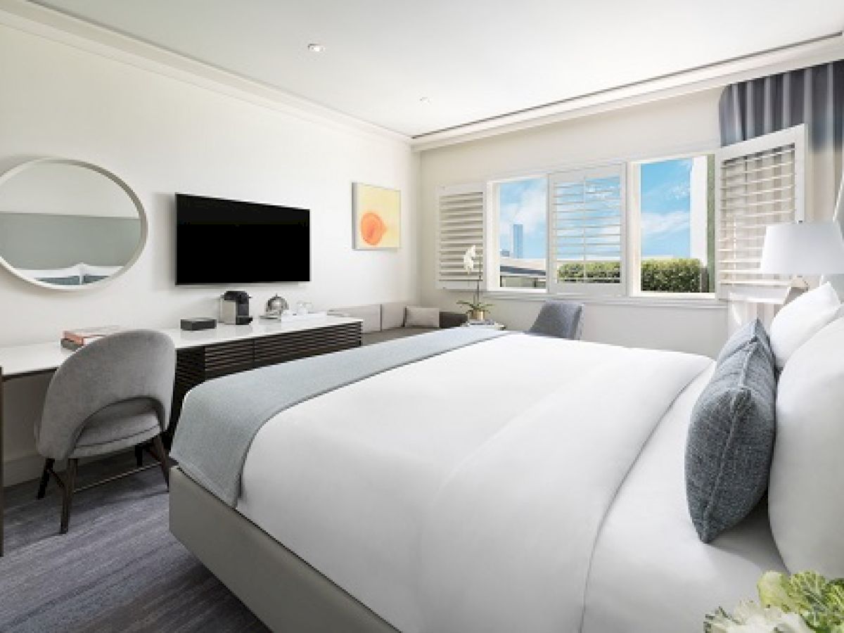 The image shows a modern hotel room with a bed, TV, desk, chair, and a window with a view. The decor is minimalist and elegant.