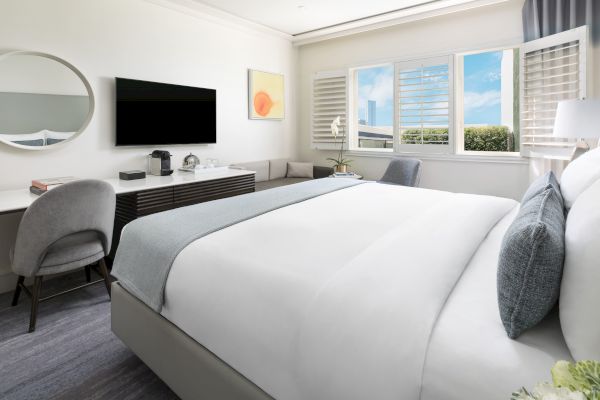 A modern hotel room with a large bed, desk, TV, and a view through a window. There's a circular mirror and minimal decor.