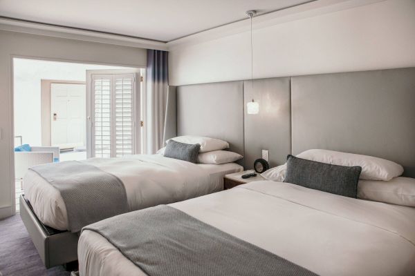 The image shows a hotel room with two neatly made beds, gray accents, a bedside table, a lamp, and an open door leading to a patio.