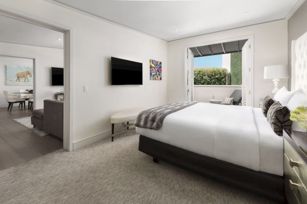 A modern bedroom with a large bed, wall-mounted TV, artwork, open patio doors, and a view of a living area with seating and a dining table.