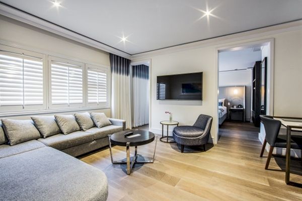 A modern living room with a gray sofa, round tables, TV, and visible bedroom, featuring light wood flooring and ceiling lights.