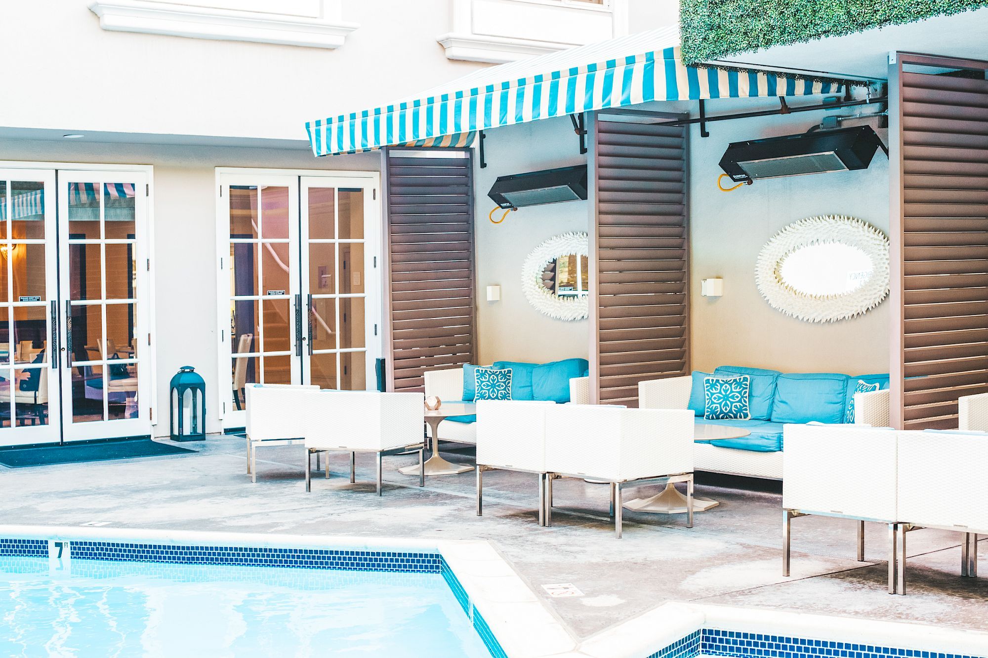 A poolside lounge area with blue and white furniture, striped awning, decorative mirrors, and French doors in the background.