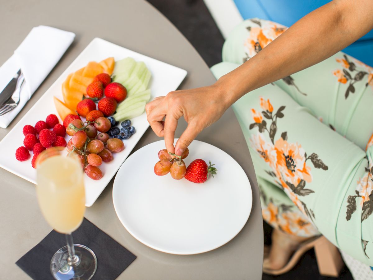 A person in floral pants picks grapes from a fruit platter with strawberries, raspberries, melons, and a drink on a table.