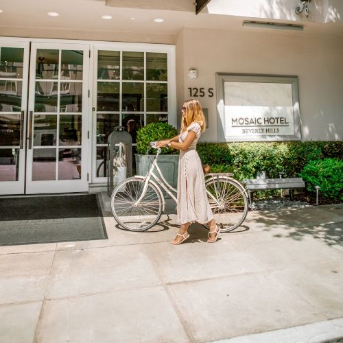 A woman walks a bicycle outside the Mosaic Hotel Beverly Hills entrance in a sunny setting.