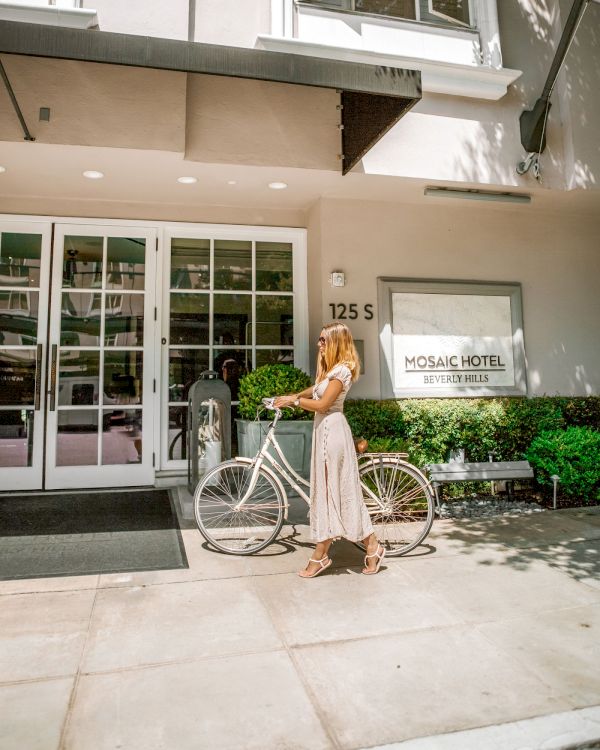 A woman walks a bicycle outside the Mosaic Hotel Beverly Hills entrance in a sunny setting.