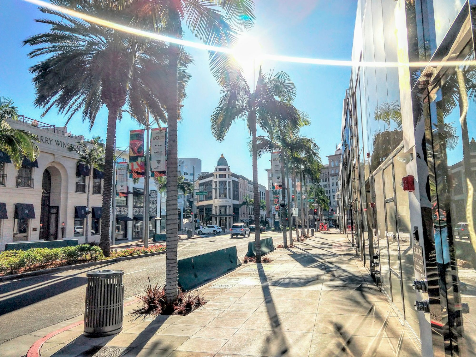 A sunny street with palm trees, storefronts, and people walking, featuring a bright sun and city buildings in the background.